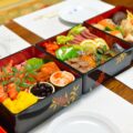 Three small osechi in black lacquer boxes filled with auspicious food and laid side by side on a table.