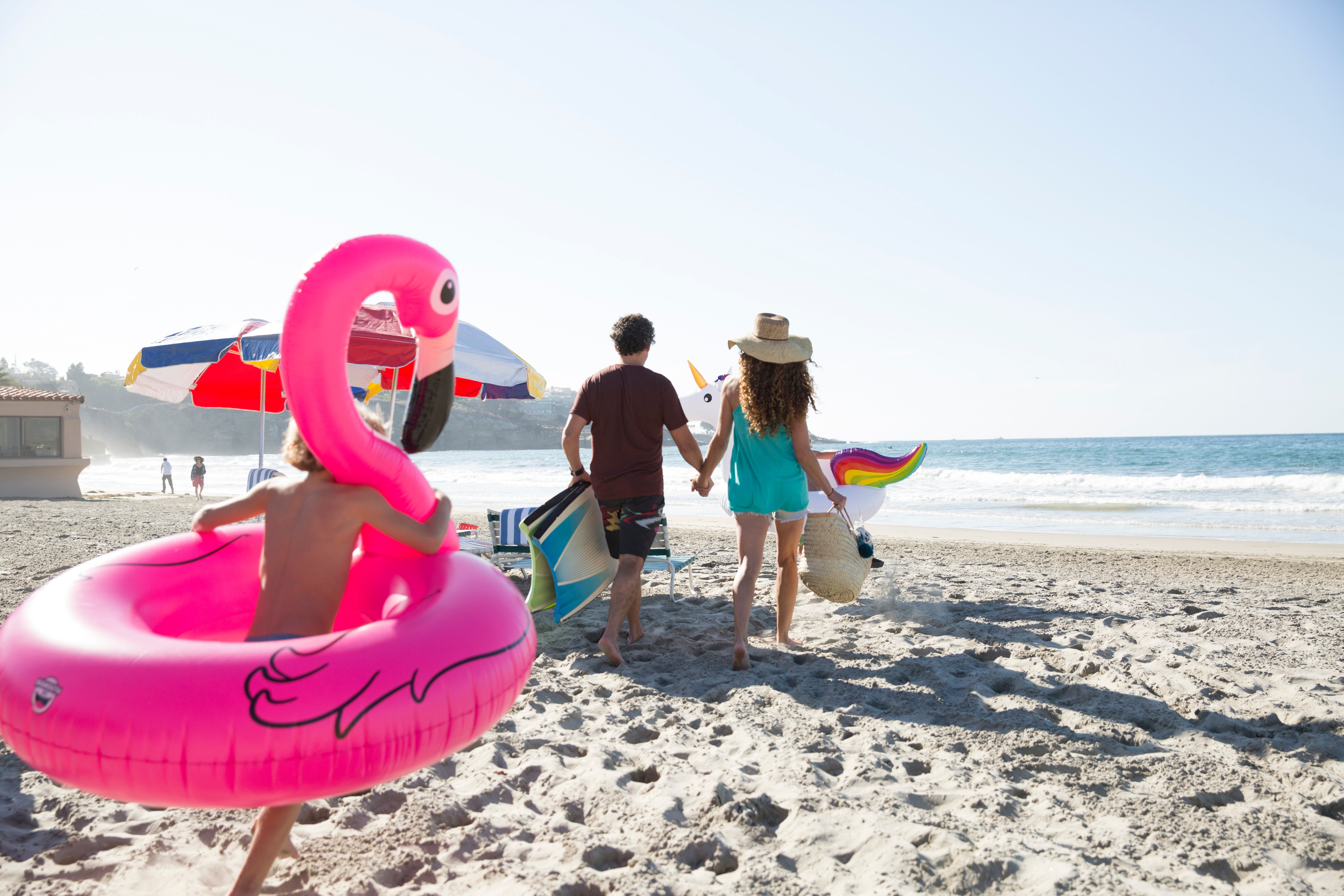 A child runs with an inflatable flamingo around him behind parents carrying beach gear on La Jolla Shores beach.