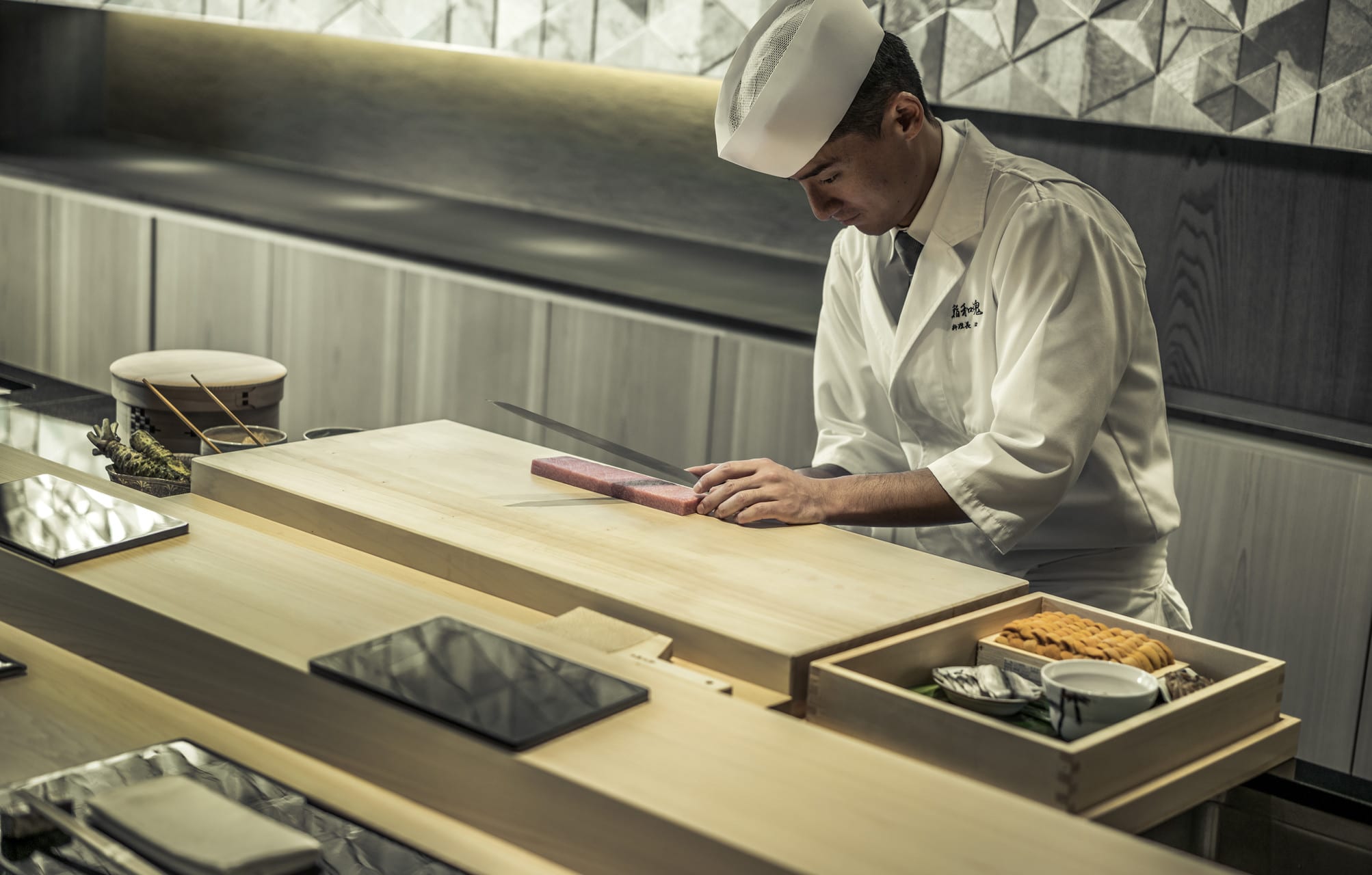 A sushi chef slices fish on a pristine wood chopping block with a long blade.