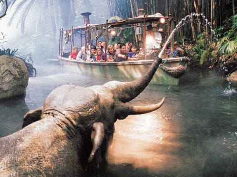 A Jungle Cruise boat passes by an elephant shooting water from its trunk.