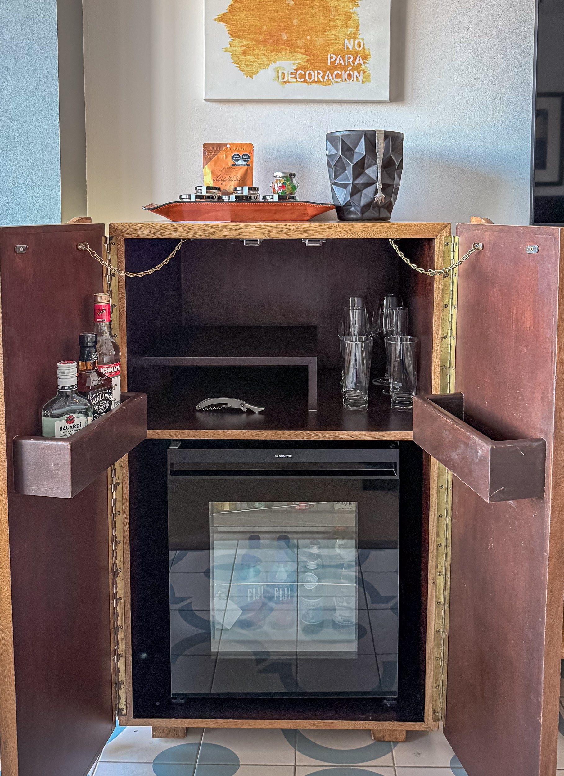 The minibar cabinet open in a guest room at The Cape, with spirits, glassware and a stocked refrigerator