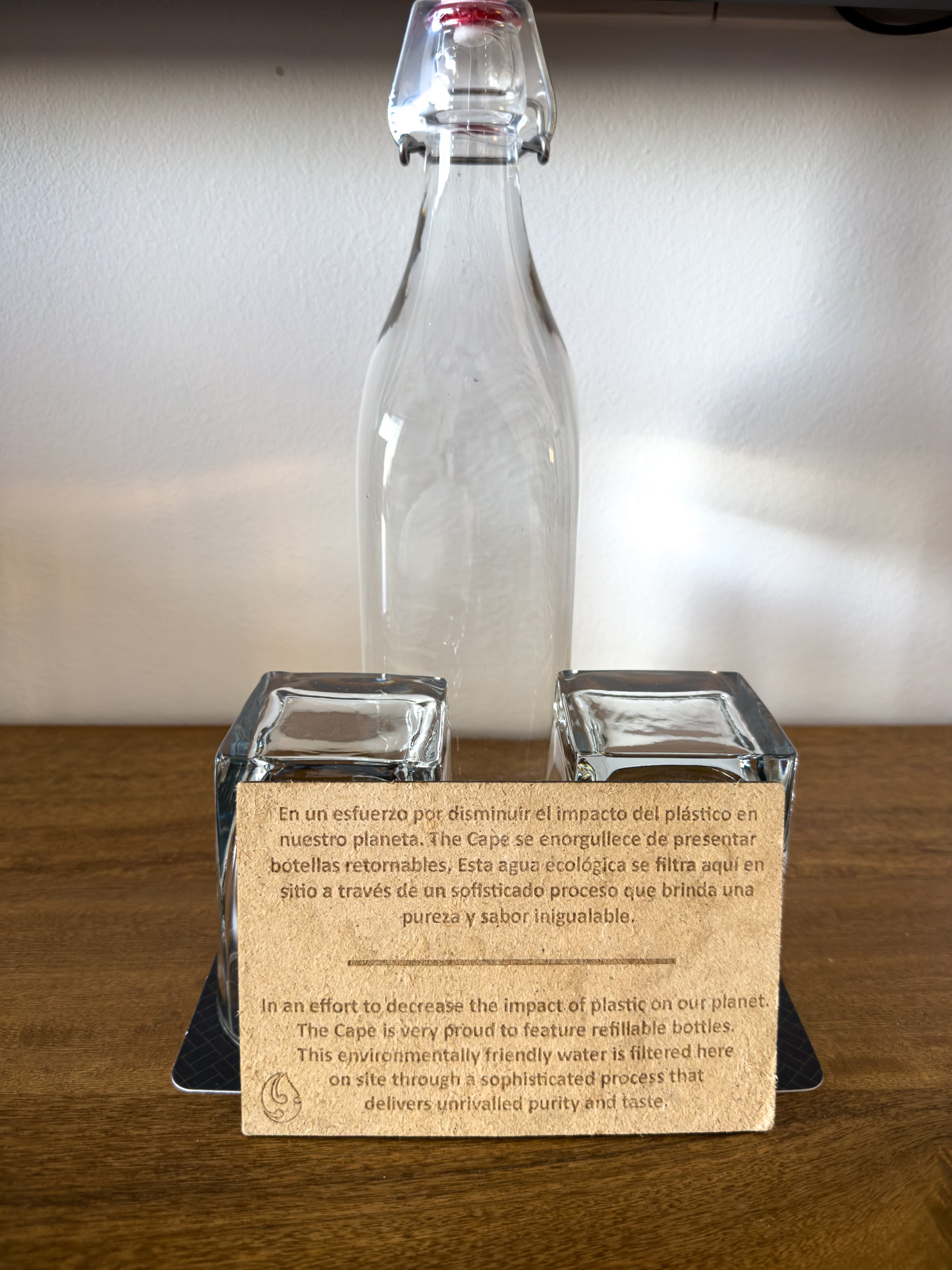 A refillable glass water bottle and tumblers in a guest room at The Cape, beside the card explaining the hotel's filtered water