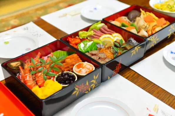 Three small osechi in black lacquer boxes filled with auspicious food and laid side by side on a table.