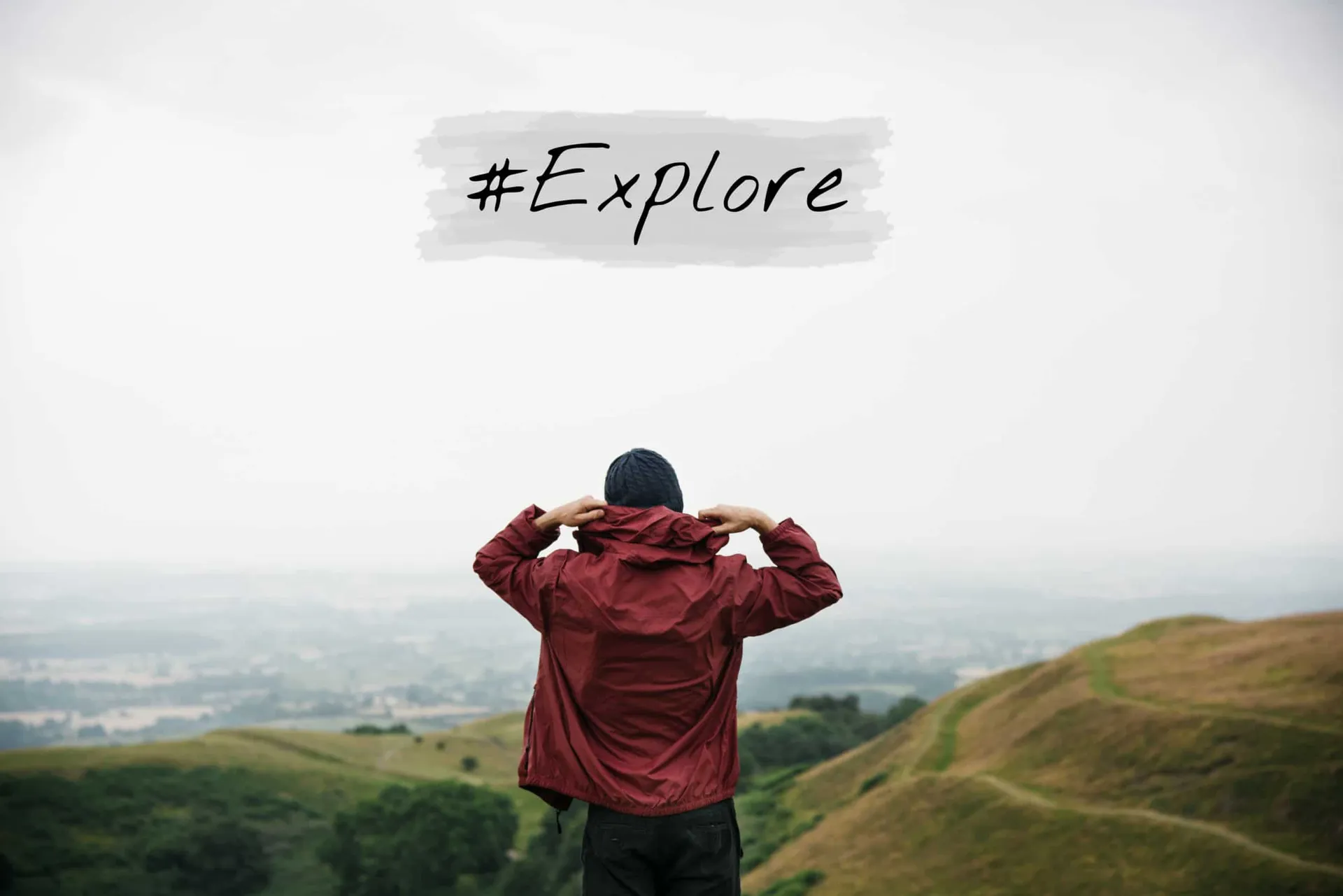 A man in a red jacket overlooks a valley with the #explore hashtag for Instagram written over the sky.