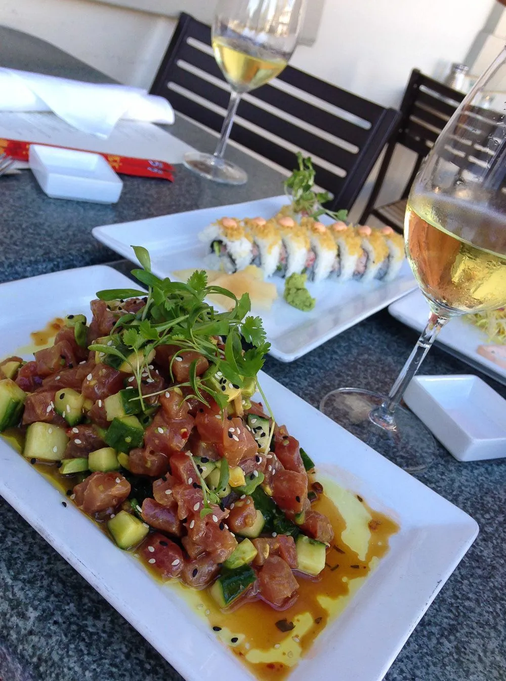 Poke and sushi on white plates near a glass of white wine at The Fishery.