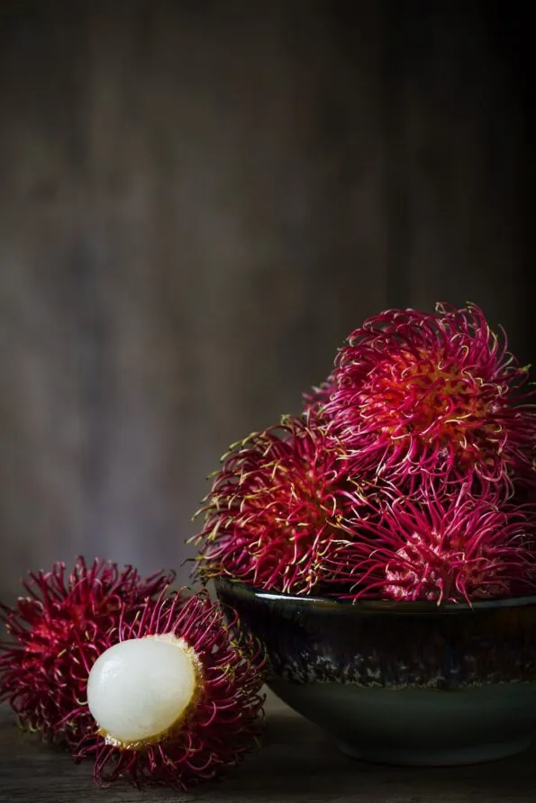 One open rambutan placed next to whole rambutans in a bowl.