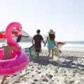 A child runs with an inflatable flamingo around him behind parents carrying beach gear on the beach.