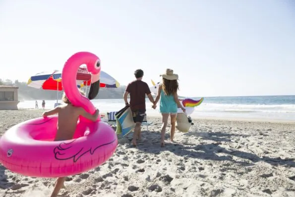 A child runs with an inflatable flamingo around him behind parents carrying beach gear on the beach.