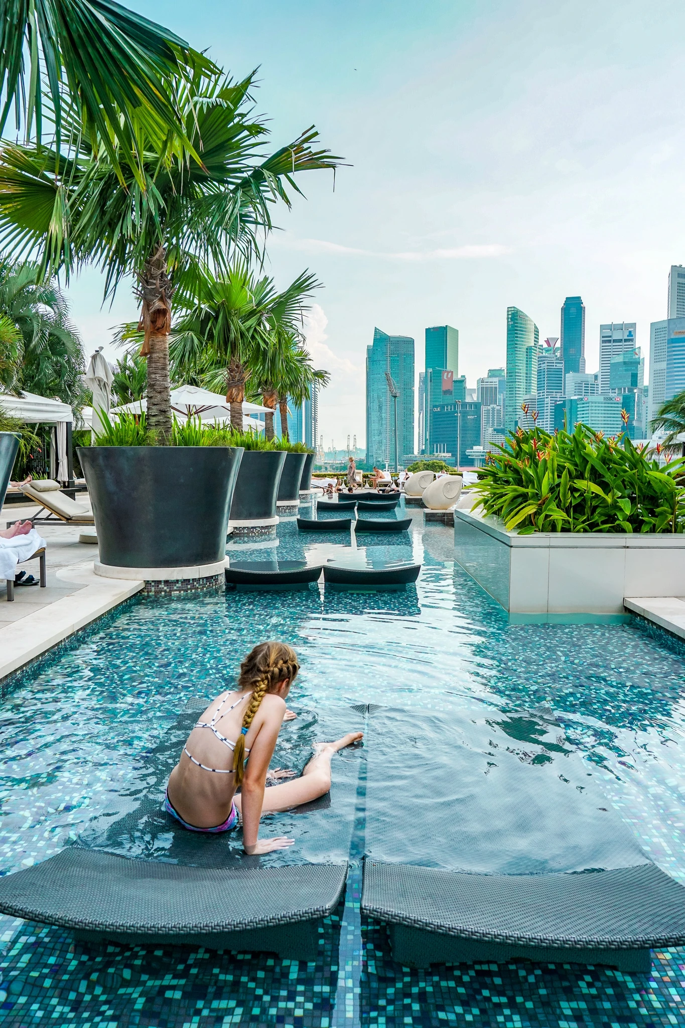 My daughter sits in a shallow pool with lounge chairs overlooking Marina Bay.