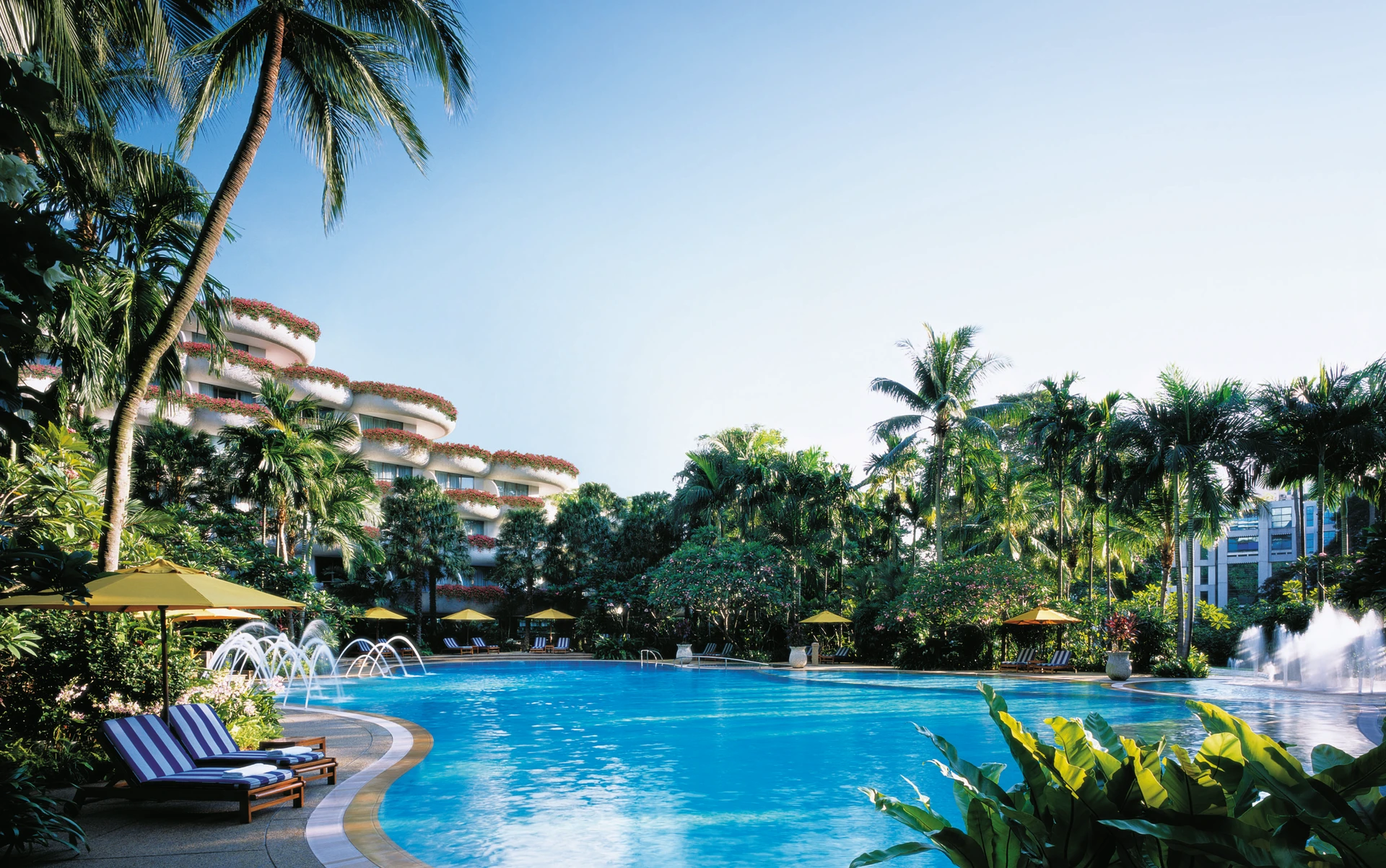Pool surrounded by tropical foliage and striped blue and white lounge chairs shaded by yellow umbrellas.
