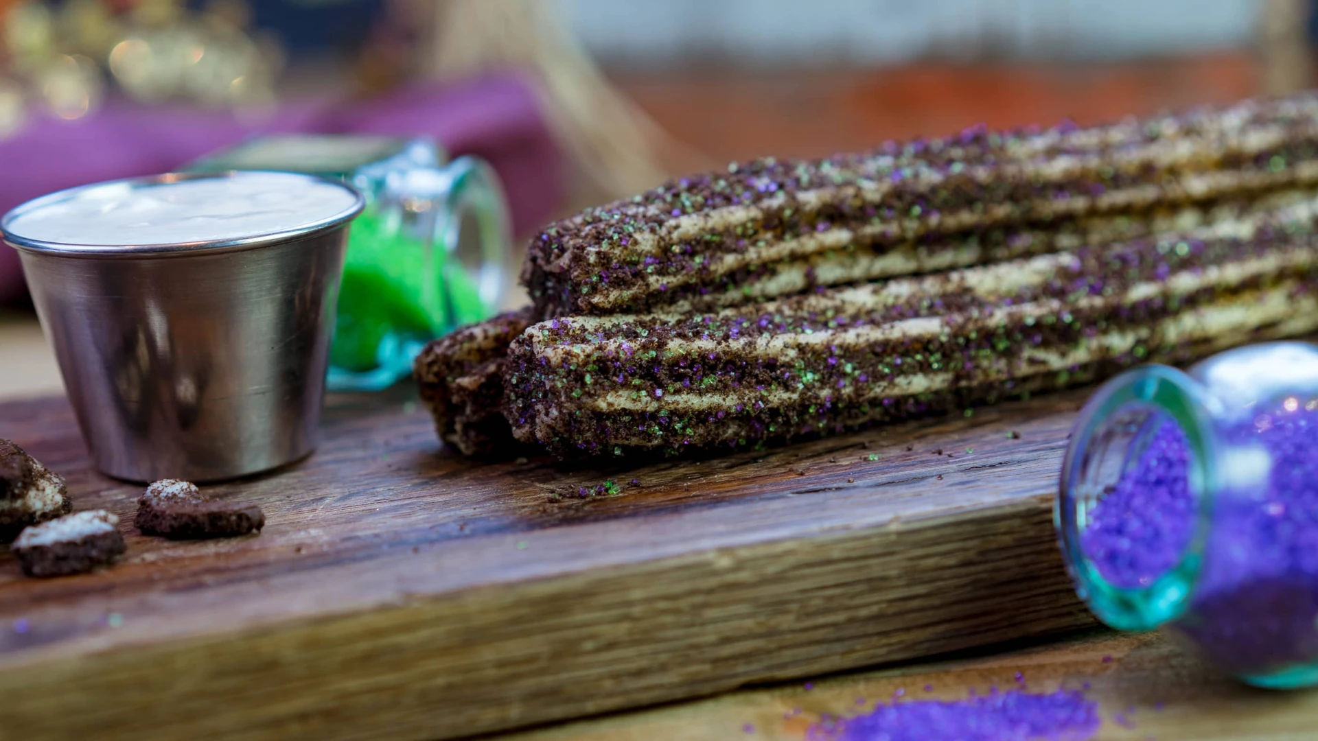 Maleficent-inspired churros resting on a wood board near a cream dipping sauce and up-ended sprinkles containers.