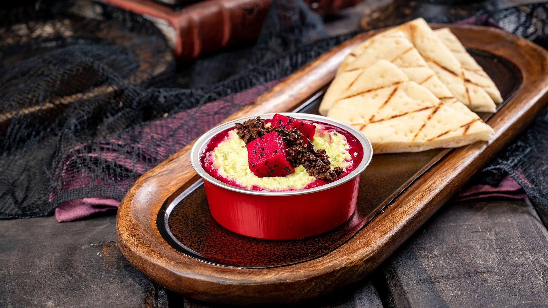 The hummus in a red bowl served on an oval platter with wedges of grilled flatbread.