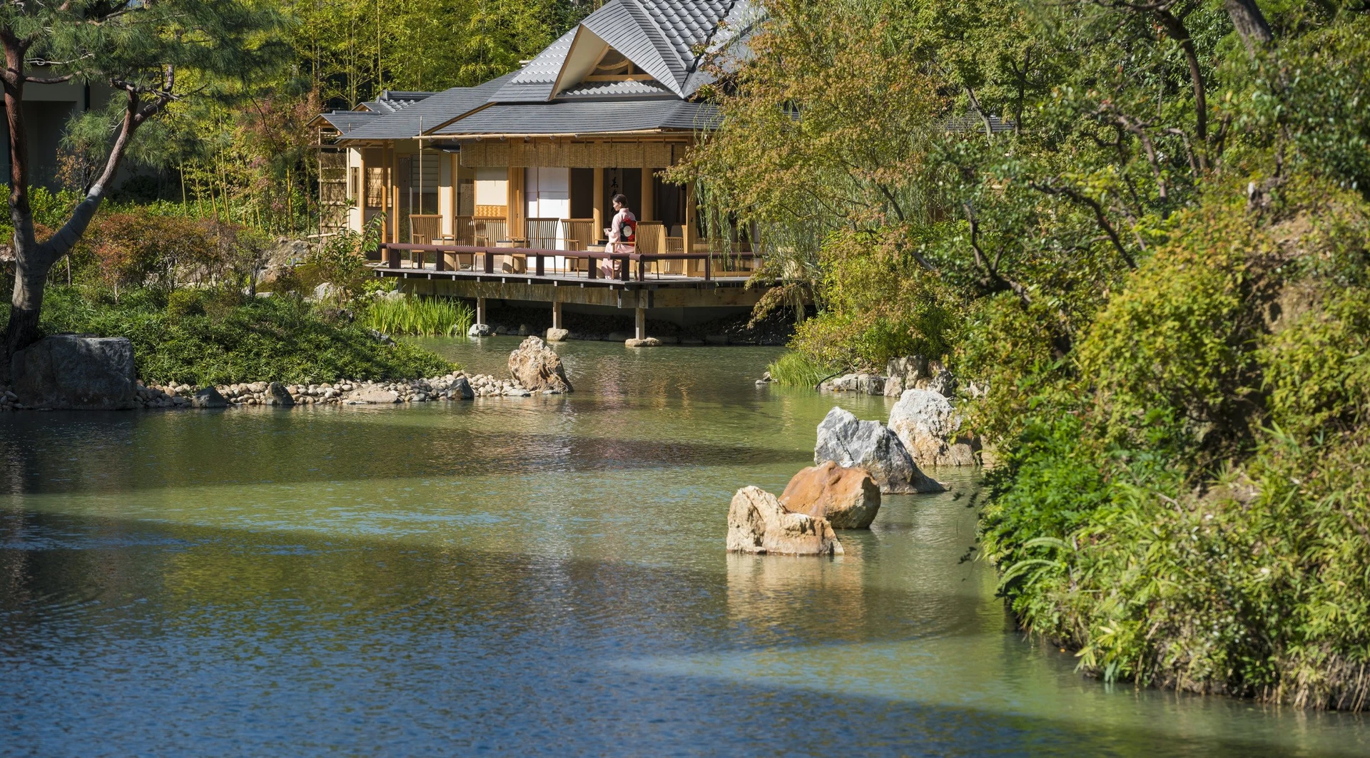 View over the water to the teahouse with a woman in a kimono walking on the terrace.