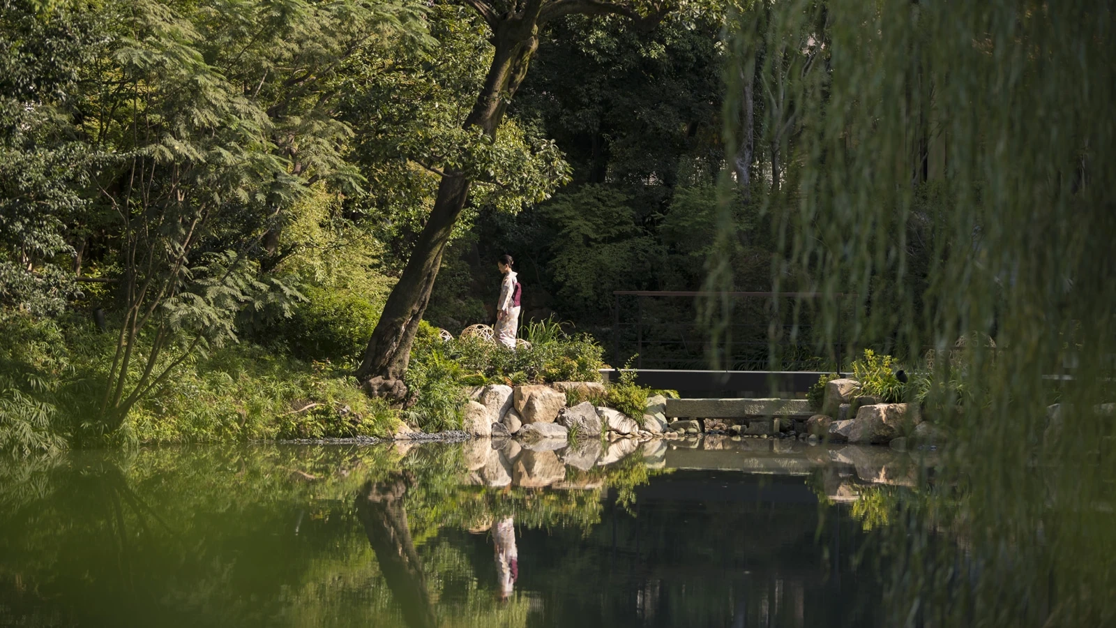 A woman in a kimono crosses a bridge over the pond in Shakusuien garden.