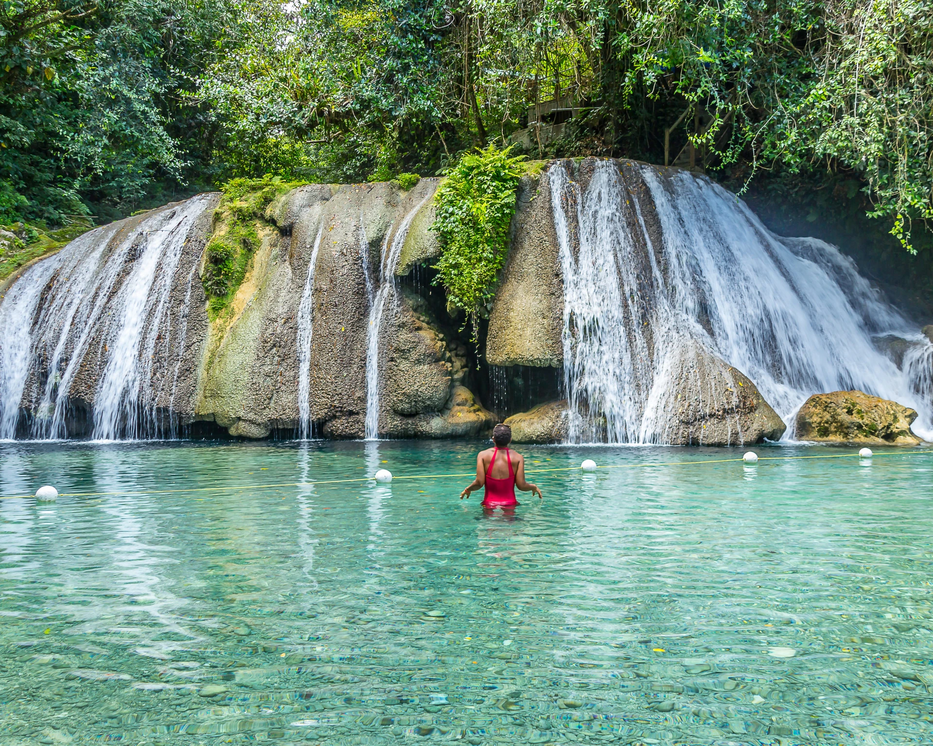 A woman in a red bathing suit wading in waist deep clear water faces Reach Falls.