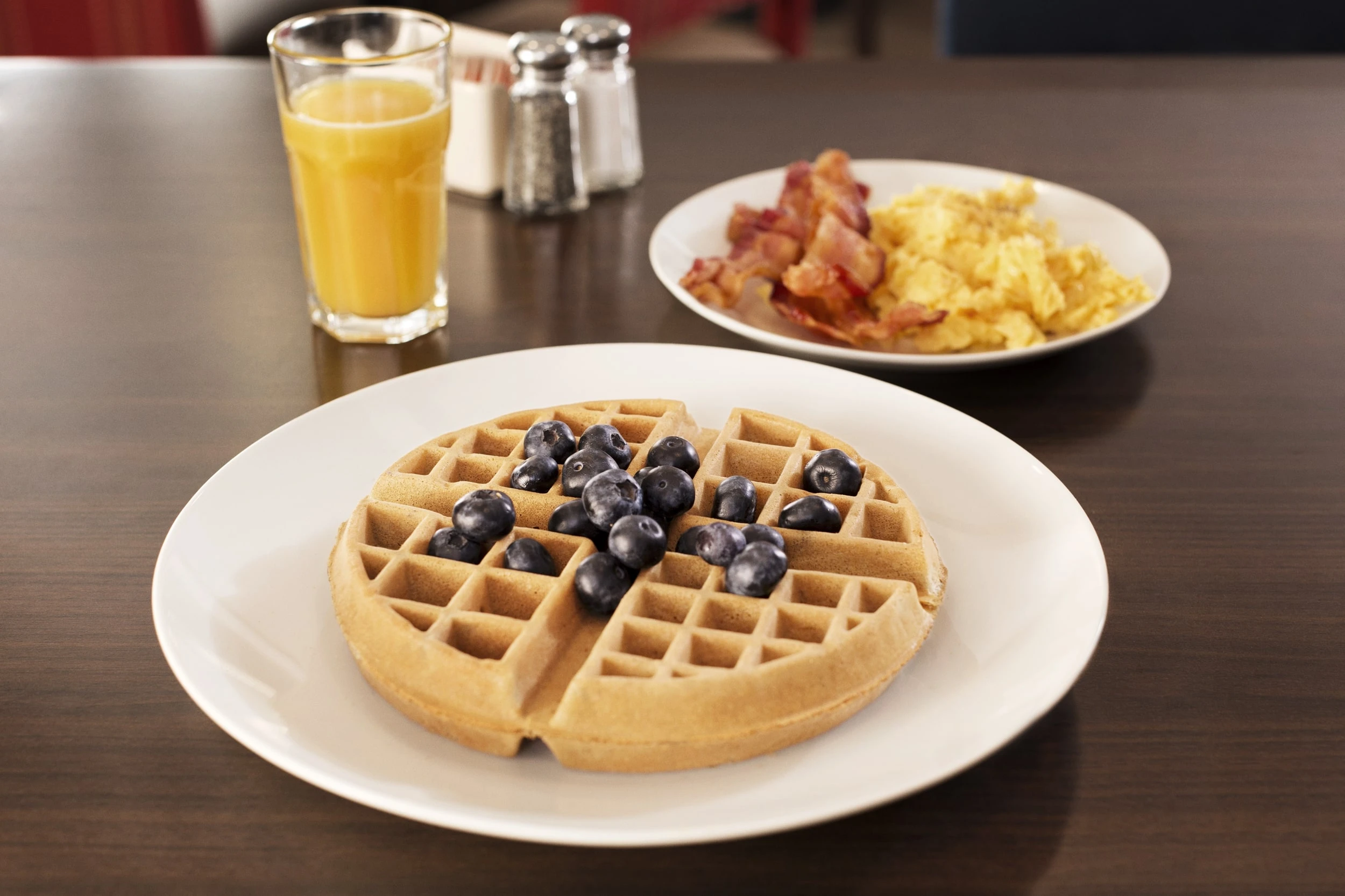 A plate of waffles next to a plate of eggs and bacon with glass of orange juice.