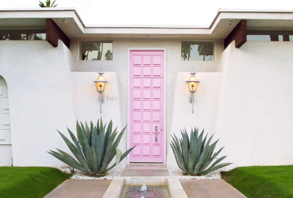 The famous cotton candy pink, narrow front door on a white one-story mid-century home, flanked by two agave succulents.