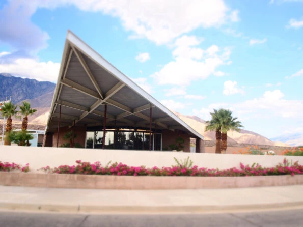 The triangular roof/overhang of the Visitor's Center juts into the sky. Mountains and palm trees are in the background.