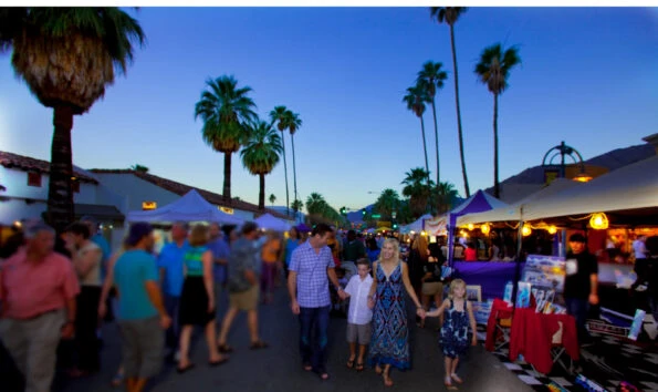 A family walks through Villagefest at dusk with vendors under white Farmer's Market-style tens on each side.