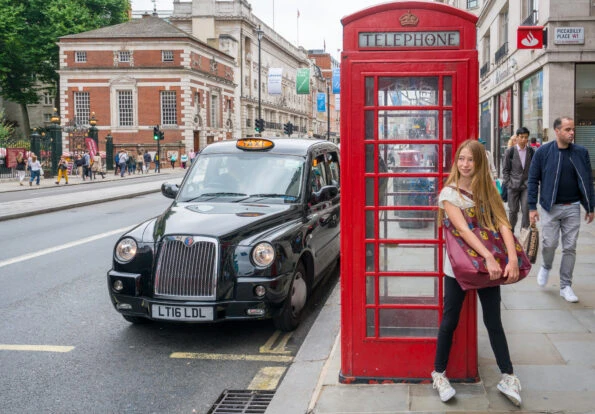 Girl in front of a red British phone booth as a traditional black taxi passes by.