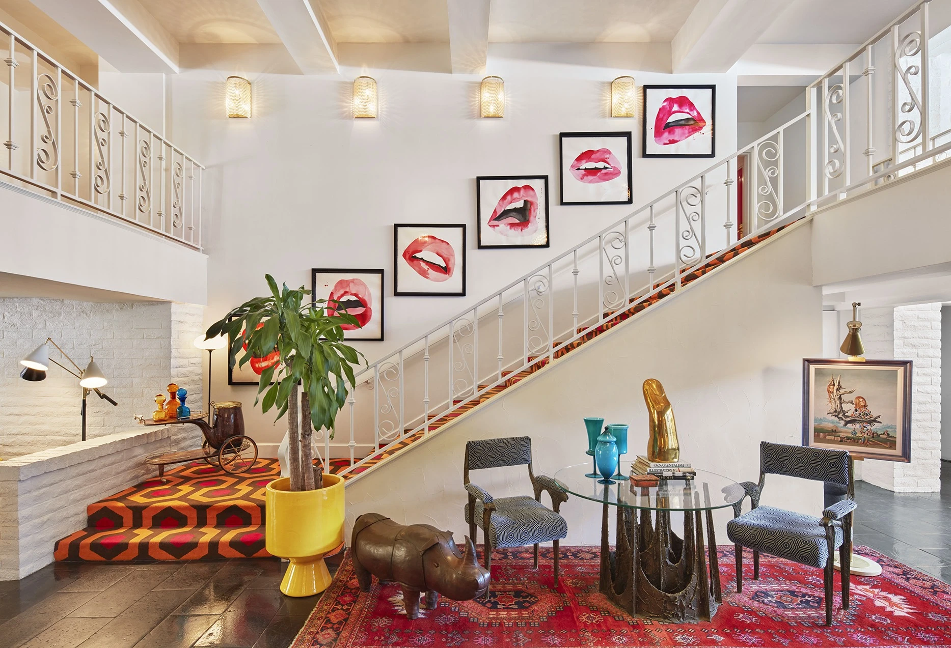 The eclectic and colorful hip lobby of the Parker Hotel with Mid-Century table and chairs, palm tree in yellow pot, metal rhino and collage of framed paintings of pink lips along a staircase.