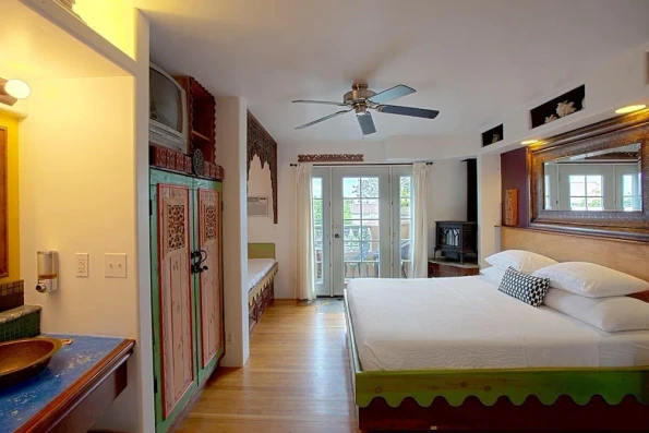 A king bedroom interior with loads of character, wood burning stove, white sheets, and Mexican-inspired colorful woodwork.