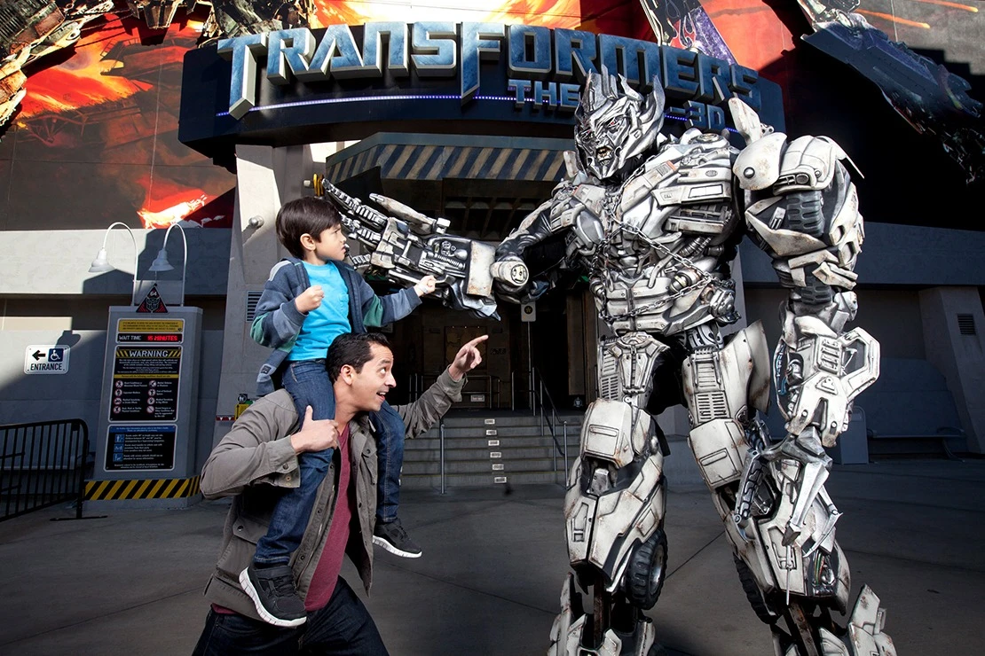A boy on a man's shoulders points his finver at Optimus Prime in front of the Transformers Ride entrance at Universal Studios Hollywood.