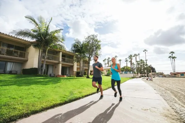 A man and woman jog down the boardwalk in front of San Diego Mission Bay Resort, near the beach.