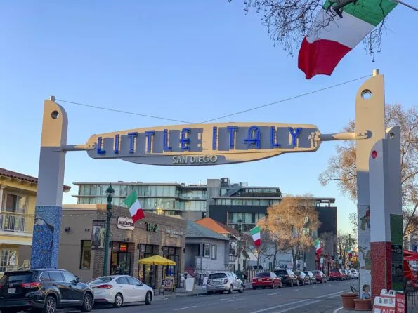 The Little Italy San Diego sign hangs over the street at golden hour with cars parked in front of restaurants and storefronts.