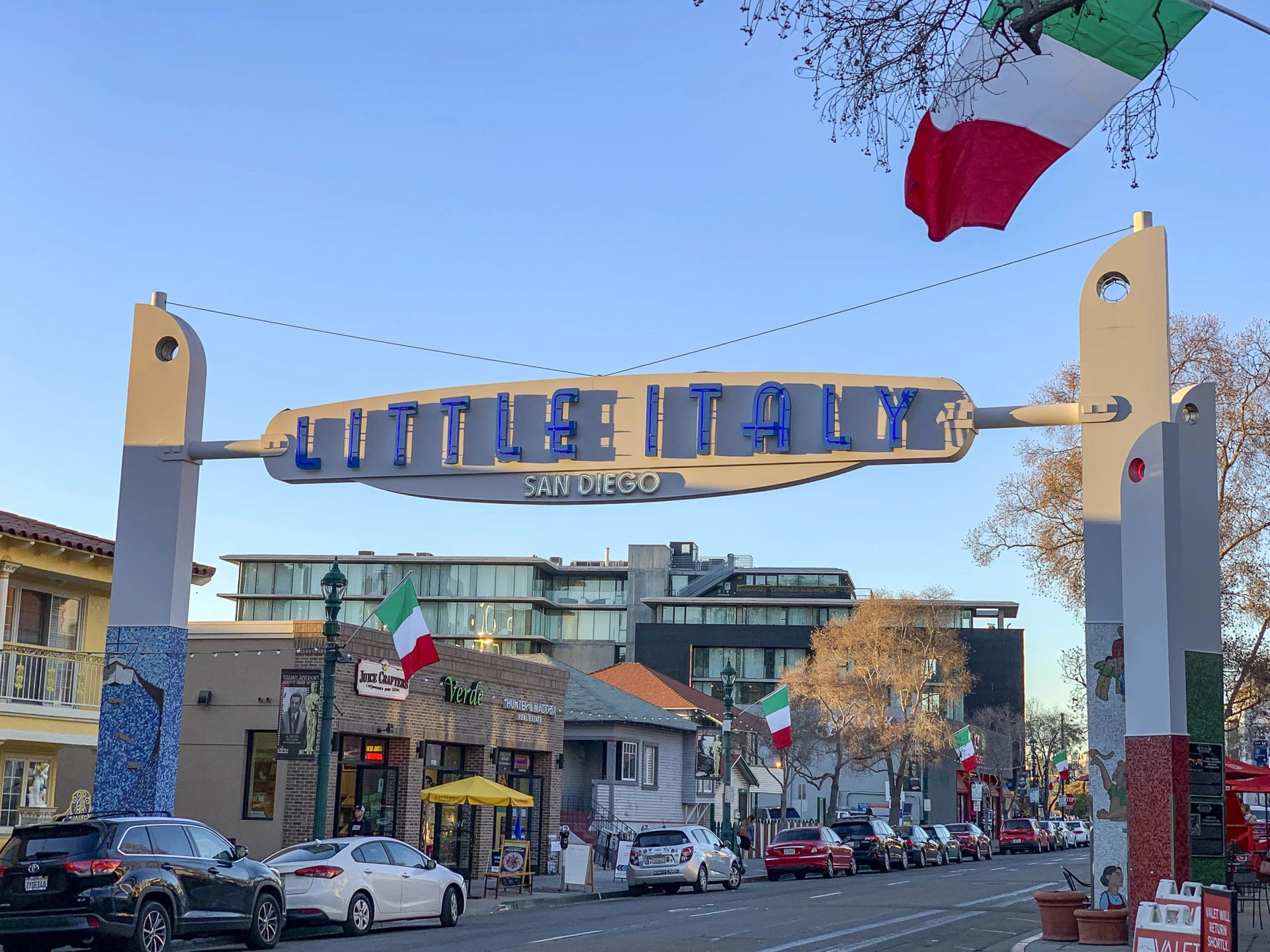 The Little Italy San Diego sign hangs over the street at golden hour with cars parked in front of restaurants and storefronts.