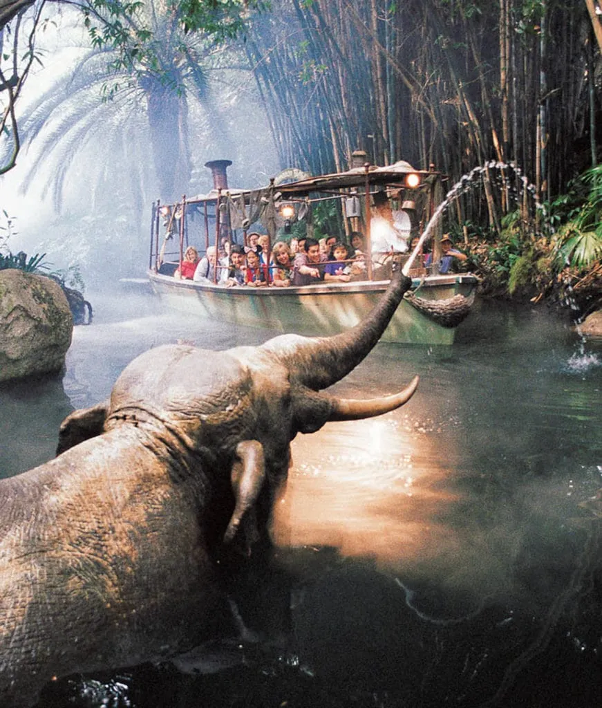 A Jungle Cruise boat passes by an elephant shooting water from its trunk.