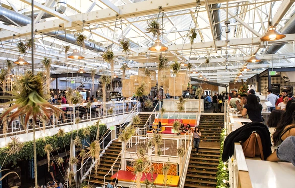 Large roof windows shed light on hanging plants in the large commercial food hall space as people dine at tables.