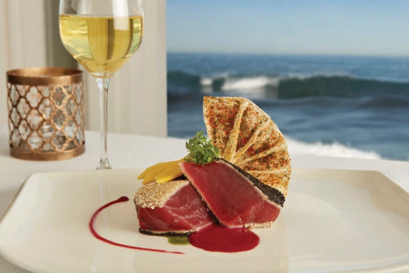 Seared ahi and a glass of white wine against the window with the ocean behind.