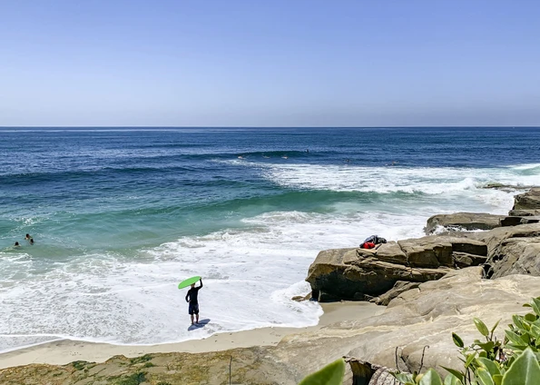 A guy stands with a green skim board on top of his head at the shoreline of Windansea Beach, La Jolla.