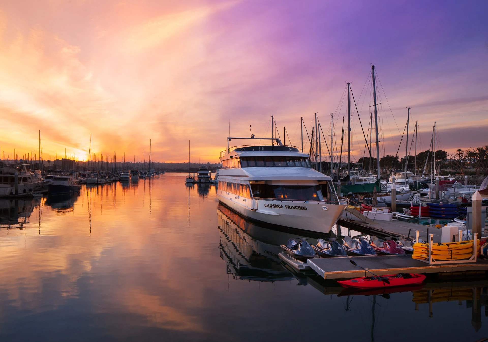 Water sports area on the bay with kayaks, jet skis, and boats at sunset.