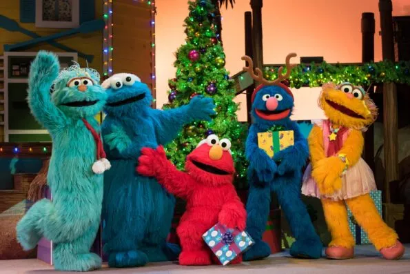 Elmo and friends hold presents in front of a Christmas tree.