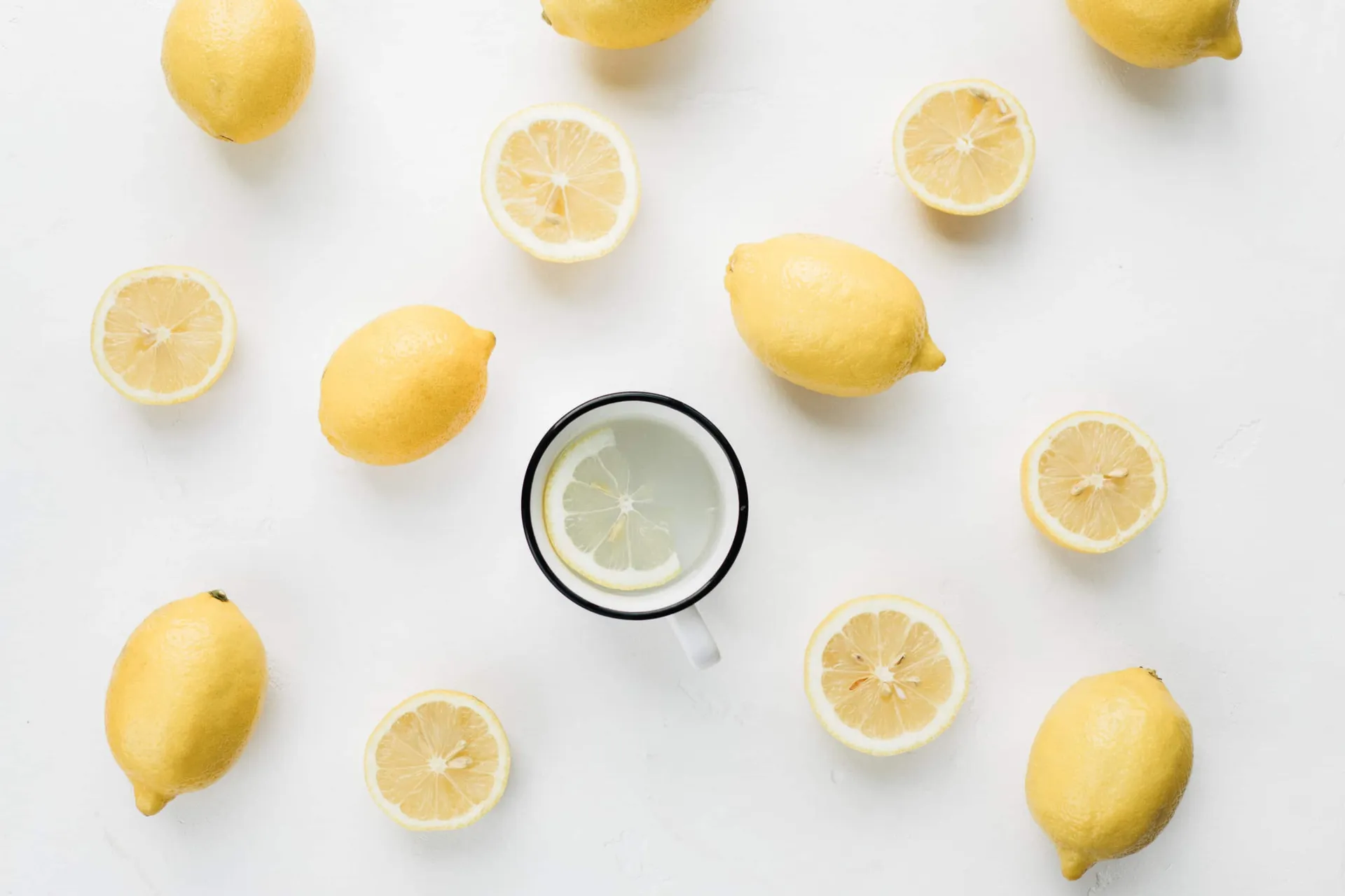 Whole and halved lemons surround a mug of warm lemon water on a table.