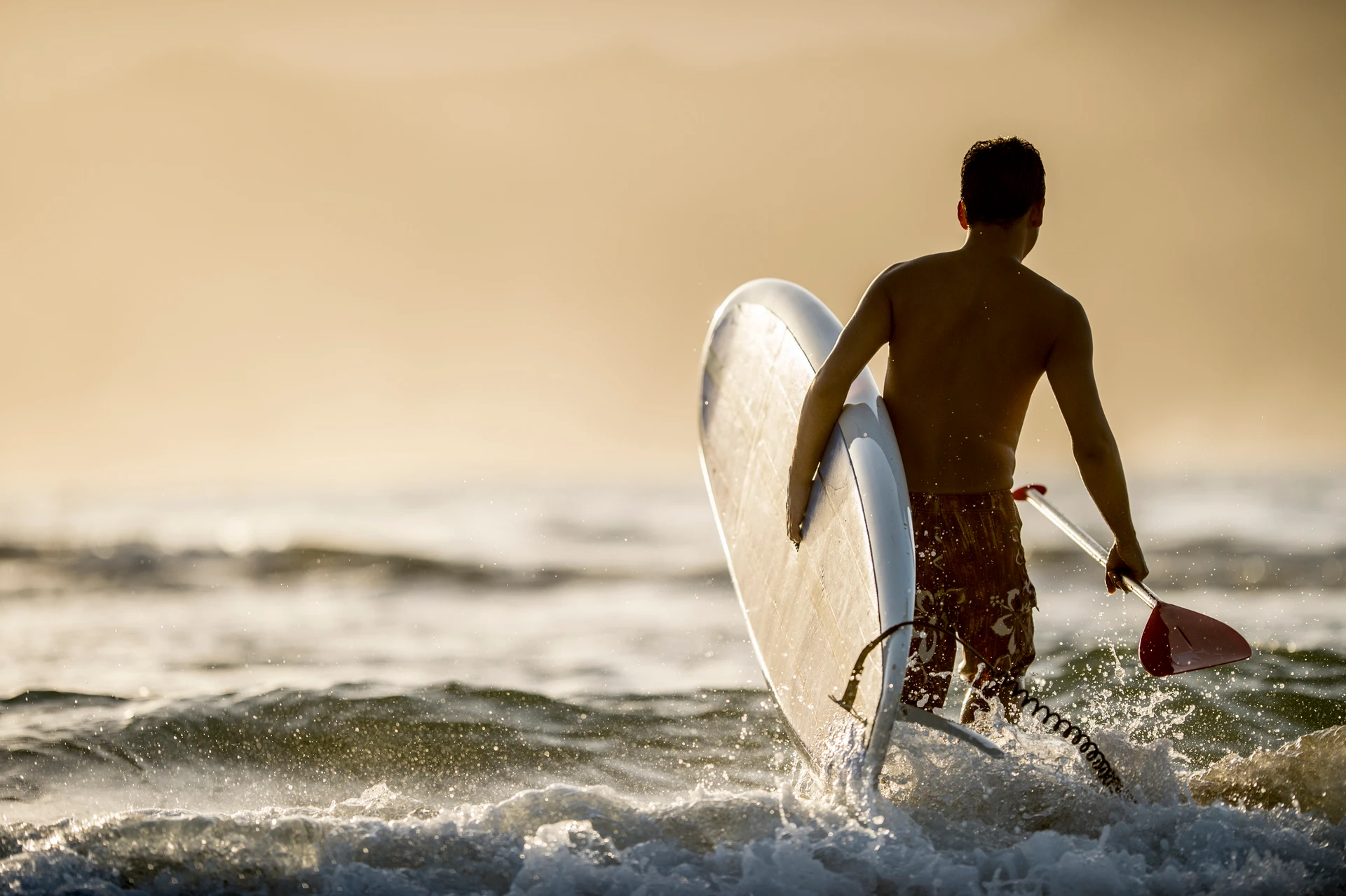 A man walks into the ocean holding a stand up paddle board and paddle.