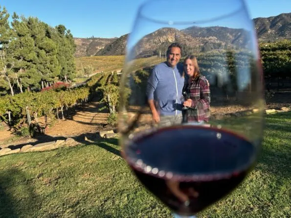 A couple stands in front of a pretty vineyard with a wine glass.