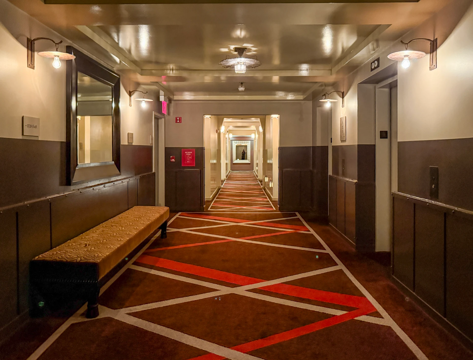 The hallway in front of the elevator with bench seating, dark woods and rich red carpet.
